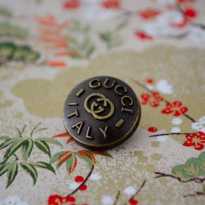 May include: A circular, antique brass button featuring the word "GUCCI" at the top and "ITALY" at the bottom, with the Gucci logo in the centre. The button rests on a patterned background with floral and foliage designs.