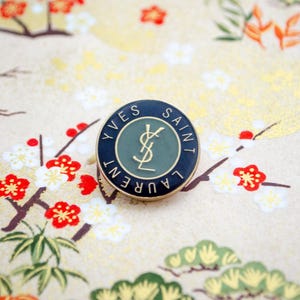 以下が含まれることがあります： イヴ・サンローランのロゴが刻まれた黒と金のピン。ロゴは緑色の背景に金色のスタイリッシュなYSLモノグラムです。