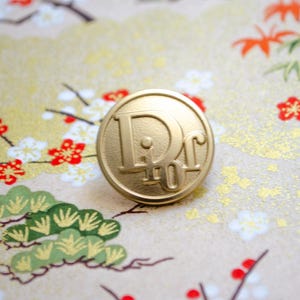 May include: A gold-colored metal button with the letters "D.C." embossed on it. The button is circular and has a slightly raised edge.