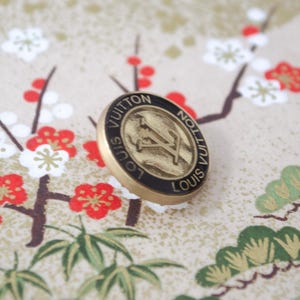 Puede incluir: Un botón Louis Vuitton dorado con borde negro, con el logotipo de la marca. El botón se encuentra sobre un fondo de un estampado floral con flores rojas y blancas y follaje verde.