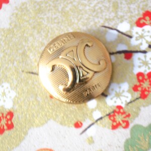 May include: A gold-coloured button with the word "CELINE" and the "C" logo embossed on the front. The button is round and has a ridged texture. The background features a floral pattern with red and white flowers.