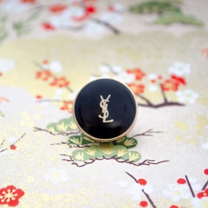 May include: A black button with a gold YSL logo on a white background with a floral pattern.