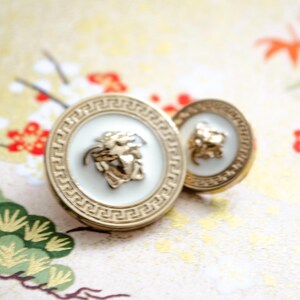 May include: Two gold-toned cufflinks with a white center and a Medusa head emblem. The cufflinks have a Greek key border design. The background is a floral pattern with red and gold accents.