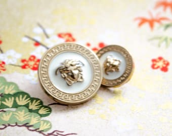 May include: Two gold-toned cufflinks with a white center and a Medusa head emblem. The cufflinks have a Greek key border design. The background is a floral pattern with red and gold accents.