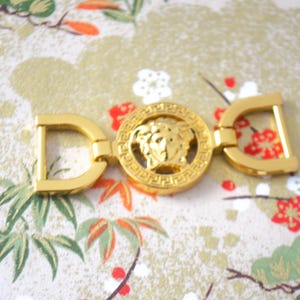 May include: Gold-toned metal buckle with a central circular medallion featuring a Medusa head design, flanked by two D-shaped elements. The buckle is set against a patterned background with floral and foliage motifs.