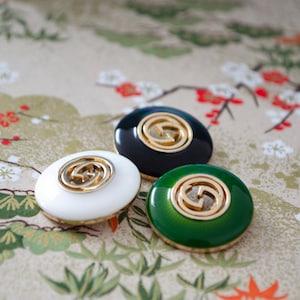 Puede incluir: Tres botones ovalados en blanco, negro y verde, cada uno con un emblema dorado. Los botones están colocados sobre un fondo estampado con motivos florales y botánicos. Los botones tienen un acabado brillante y pulido.