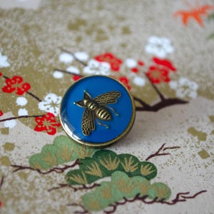 May include: A circular button featuring a smooth, deep blue centre inlay. A metallic, bronze-toned bee with detailed wing patterns is centred on the blue surface. The button has a raised, dark bronze-coloured metal rim surrounding the blue inlay.