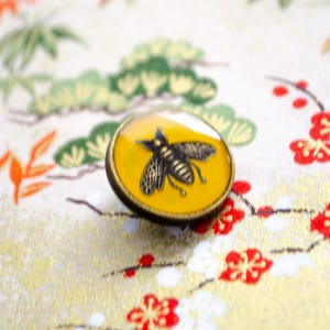 May include: A circular button featuring a yellow resin center with a detailed black bee illustration. The button has a metallic bronze-toned rim and a smooth, slightly domed surface, showcasing a clear contrast between the insect graphic and the background.
