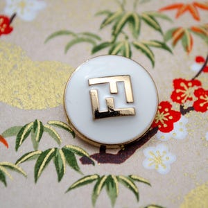 May include: White round button with a gold interlocking 'F' logo.