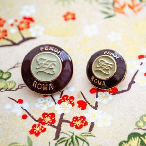 May include: A pair of Fendi Roma earrings. The earrings are circular with a dark brown border and a pale green centre. The centre features the Fendi logo in gold. The word "FENDI" is at the top and "ROMA" at the bottom in gold lettering.