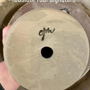 Puede incluir: Base de maceta de cerámica de color claro con una firma oscura y un orificio de drenaje. El texto "Custom Your Signature" es visible encima de la maceta. La maceta tiene una superficie moteada y texturizada.