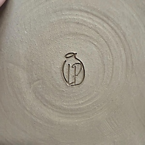May include: Close-up of the bottom of a handmade ceramic plate. The plate is a light beige color with a textured surface. The artist's initials, "LP", are carved into the center within a stylized design.