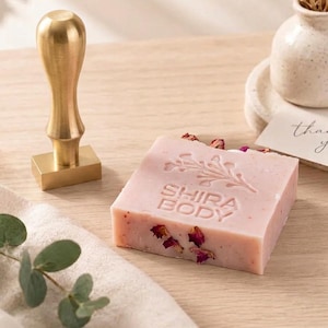 Peut inclure: Un savon carré rose clair incrusté de pétales de fleurs séchées, portant le texte en relief SHIRA BODY et un motif de branche fleurie. À côté du savon se trouve un tampon rectangulaire en laiton poli avec un manche lisse et arrondi.