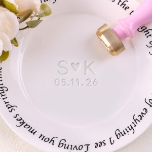 Puede incluir: Plato de cerámica blanco con las iniciales en relieve "S♡K" y la fecha "05.11.26". Un sello de cera rosa y dorado descansa sobre el plato. Arreglo floral en la esquina superior izquierda. Escritura negra rodea el borde.