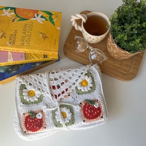 Puede incluir: Bolso de crochet blanco con un patr&oacute;n de fresas y margaritas. El bolso est&aacute; doblado y colocado sobre una superficie blanca. Hay libros y una taza de t&eacute; en el fondo.