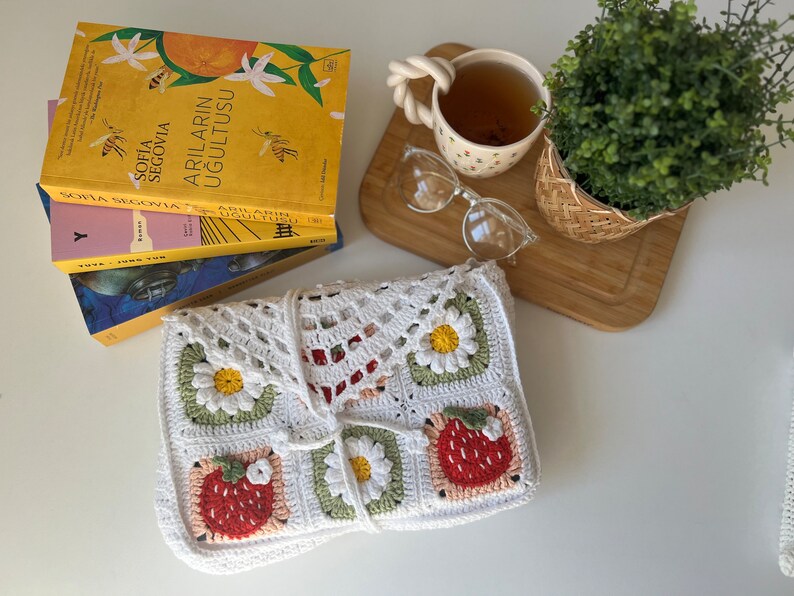 Puede incluir: Bolso de crochet blanco con un patr&oacute;n de fresas y margaritas. El bolso est&aacute; doblado y colocado sobre una superficie blanca. Hay libros y una taza de t&eacute; en el fondo.