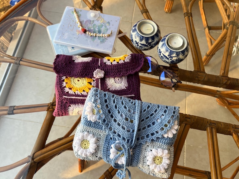 May include: Two crocheted bags, one purple with yellow and white flowers and one blue with white flowers, on a wicker table with candles and glasses.