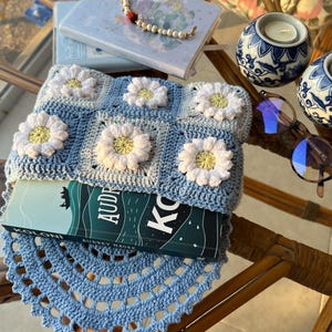 May include: A blue crocheted book cover with a daisy pattern. The cover is on top of a book titled "Koloni" by Audrey Magee. The book is on a blue crocheted doily.