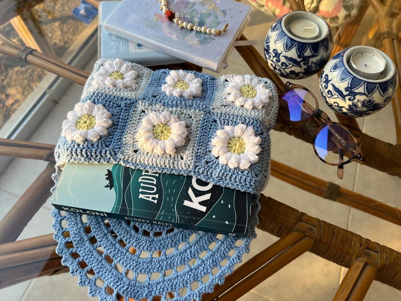 May include: A blue crocheted book cover with a daisy pattern. The cover is on top of a book titled "Koloni" by Audrey Magee. The book is on a blue crocheted doily.