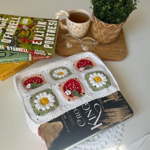 Puede incluir: Una bolsa de libro de crochet blanco con un patr&oacute;n de fresa y margarita. La bolsa contiene un libro titulado "Leigh Cro King".