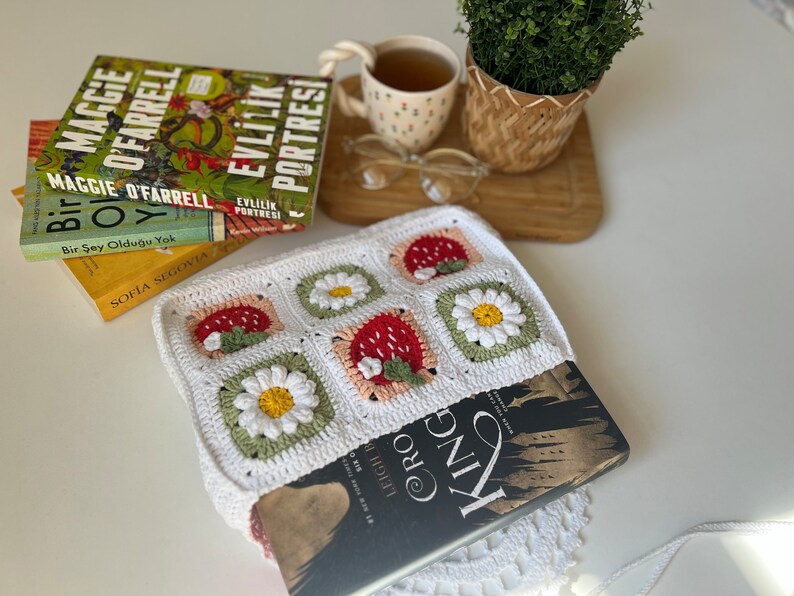 Puede incluir: Una bolsa de libro de crochet blanco con un patr&oacute;n de fresa y margarita. La bolsa contiene un libro titulado "Leigh Cro King".