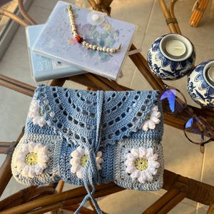 May include: A blue crocheted clutch bag with white daisy flowers. The bag has a drawstring closure and a long blue cord.