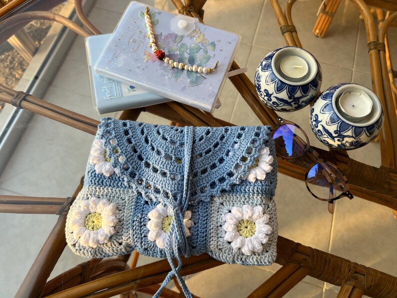 May include: A blue crocheted clutch bag with white daisy flowers. The bag has a drawstring closure and a long blue cord.