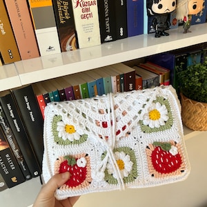 Puede incluir: Una bolsa de crochet blanca con un dise&ntilde;o floral. La bolsa tiene un patr&oacute;n de margarita, fresa y hoja. La bolsa est&aacute; atada con una cuerda blanca.