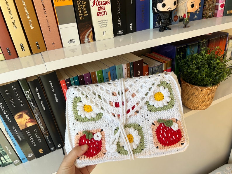Puede incluir: Una bolsa de crochet blanca con un dise&ntilde;o floral. La bolsa tiene un patr&oacute;n de margarita, fresa y hoja. La bolsa est&aacute; atada con una cuerda blanca.