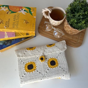 Puede incluir: Una bolsa blanca de ganchillo con un patr&oacute;n de girasol y un bot&oacute;n amarillo. La bolsa est&aacute; sobre una superficie blanca junto a una pila de libros y una taza de t&eacute;.