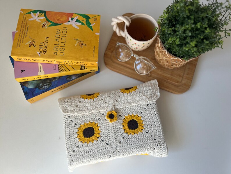 Puede incluir: Una bolsa blanca de ganchillo con un patr&oacute;n de girasol y un bot&oacute;n amarillo. La bolsa est&aacute; sobre una superficie blanca junto a una pila de libros y una taza de t&eacute;.