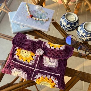 May include: A crocheted purple and pink clutch with a floral design. The clutch has a flap closure and a small flower embellishment. The clutch is sitting on a table with a pair of glasses and two decorative candle holders.