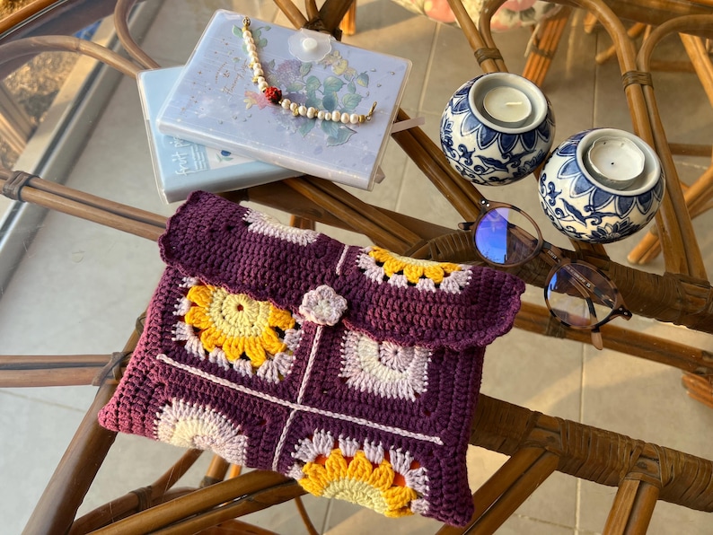 May include: A crocheted purple and pink clutch with a floral design. The clutch has a flap closure and a small flower embellishment. The clutch is sitting on a table with a pair of glasses and two decorative candle holders.
