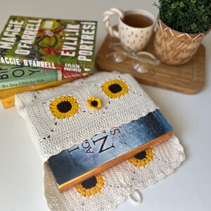 Puede incluir: Una funda de libro de crochet blanco con un dise&ntilde;o de girasol. La funda contiene un libro con una cubierta azul y el t&iacute;tulo "The Age of Z" visible.