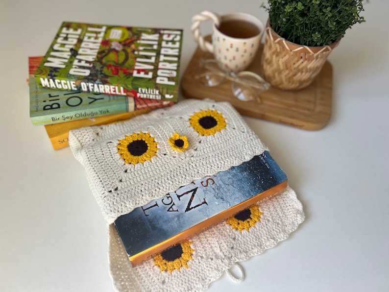 Puede incluir: Una funda de libro de crochet blanco con un dise&ntilde;o de girasol. La funda contiene un libro con una cubierta azul y el t&iacute;tulo "The Age of Z" visible.