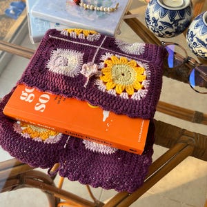 May include: A purple crocheted book cover with a floral design featuring yellow, pink, and white flowers. The cover is wrapped around a book titled "Go So" by Claire Daverley.