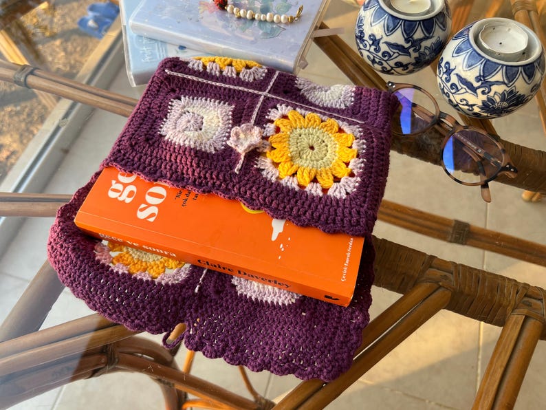 May include: A purple crocheted book cover with a floral design featuring yellow, pink, and white flowers. The cover is wrapped around a book titled "Go So" by Claire Daverley.