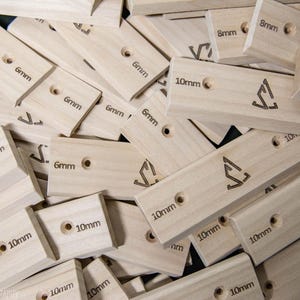 May include: A pile of light-colored wooden climbing holds. Each hold has a hole and is labeled with measurements like "6mm", "8mm", and "10mm". A logo is etched on some of the holds. The holds are rectangular and appear to be new.