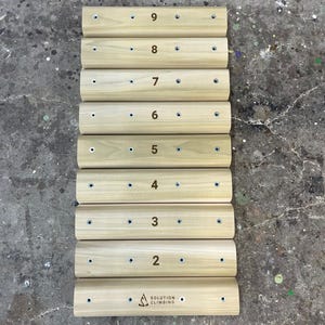 May include: A set of nine wooden climbing holds, numbered 2 through 9, with a Solution Climbing logo on the bottom hold. The holds are arranged vertically and have a light brown color.