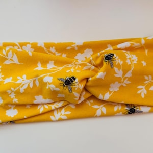 Puede incluir: Diadema amarilla con un patrón floral blanco y abejas. La diadema está retorcida y anudada.