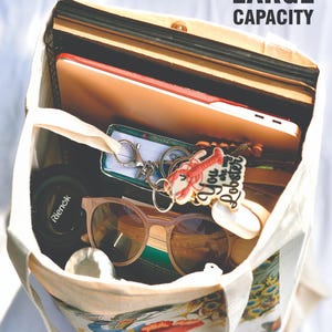 Peut inclure: Un sac fourre-tout en toile beige rempli de divers objets. &Agrave; l'int&eacute;rieur se trouvent une tablette, des livres, des lunettes de soleil, un porte-cl&eacute;s avec un motif de homard et d'autres effets personnels. Le sac a un imprim&eacute; paon et floral en bas et le texte "LARGE CAPACITY" est visible.