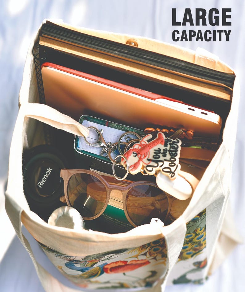 Peut inclure: Un sac fourre-tout en toile beige rempli de divers objets. &Agrave; l'int&eacute;rieur se trouvent une tablette, des livres, des lunettes de soleil, un porte-cl&eacute;s avec un motif de homard et d'autres effets personnels. Le sac a un imprim&eacute; paon et floral en bas et le texte "LARGE CAPACITY" est visible.