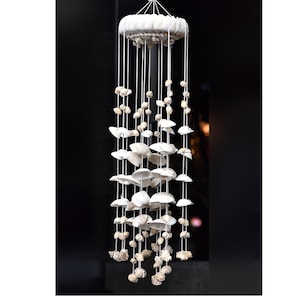 Puede incluir: Un carillón de conchas blancas con varios niveles de conchas colgando de una base circular blanca. Las conchas están dispuestas en cascada, creando un aspecto delicado y aireado.