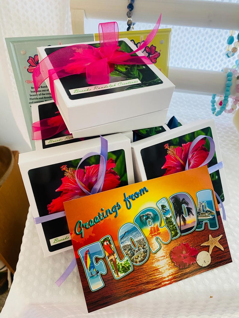 Pode incluir: Uma pilha de pequenas caixas de presente brancas com fita rosa e roxa. As caixas s&atilde;o decoradas com uma flor de hibisco e o texto "Brecks Randolph Creations". Um cart&atilde;o postal com o texto "Greetings from FLORIDA" est&aacute; em primeiro plano.