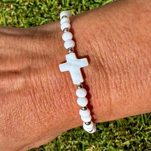 May include: A stretch bracelet featuring small white spherical beads and metallic gold-tone spacer beads, centered with a white cross-shaped bead. The design alternates between the white spheres and the gold-tone accents throughout the band.