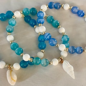 May include: Three beaded bracelets with blue, white, and gold accents. The bracelets feature blue and white beads, with gold-colored spacers. Each bracelet has a seashell charm. The bracelets are on a white surface.