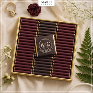 Könnte beinhalten: Eine Hochzeitsgeschenkbox mit goldenem Rahmen, gefüllt mit weinroten und dunkelbraunen Schokoladenstangen. Eine Schokoladentafel mit goldener Aufschrift "The Wedding of A&G" liegt obenauf. Ein goldener Ring und eine weinrote Rose sind ebenfalls zu sehen.