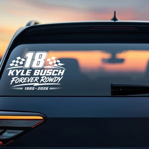 May include: White decal on a car's rear window. The decal features the number 18, a checkered flag design, and the text "KYLE BUSCH FOREVER ROWDY 1985-2026". The car is dark blue, with a sunset visible through the window.
