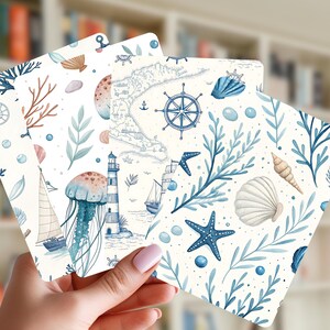 May include: A hand holds three square cards with nautical-themed illustrations. The cards feature watercolor designs of seashells, starfish, a lighthouse, a map, and a ship, in shades of blue and beige.