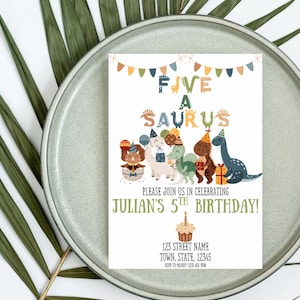 Op de afbeelding: Een verjaardagsuitnodiging met de tekst "FIVE A SAURUS" en "JULIAN'S 5TH BIRTHDAY!". Het bevat cartoon dinosaurussen en een cupcake illustratie. De uitnodiging staat op een witte achtergrond met een grijze plaat en groene bladeren.